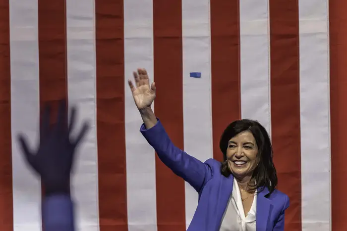 Gov. Kathy Hochul, in a blue suit jacket, waves to supporters against a backdrop of the American flag